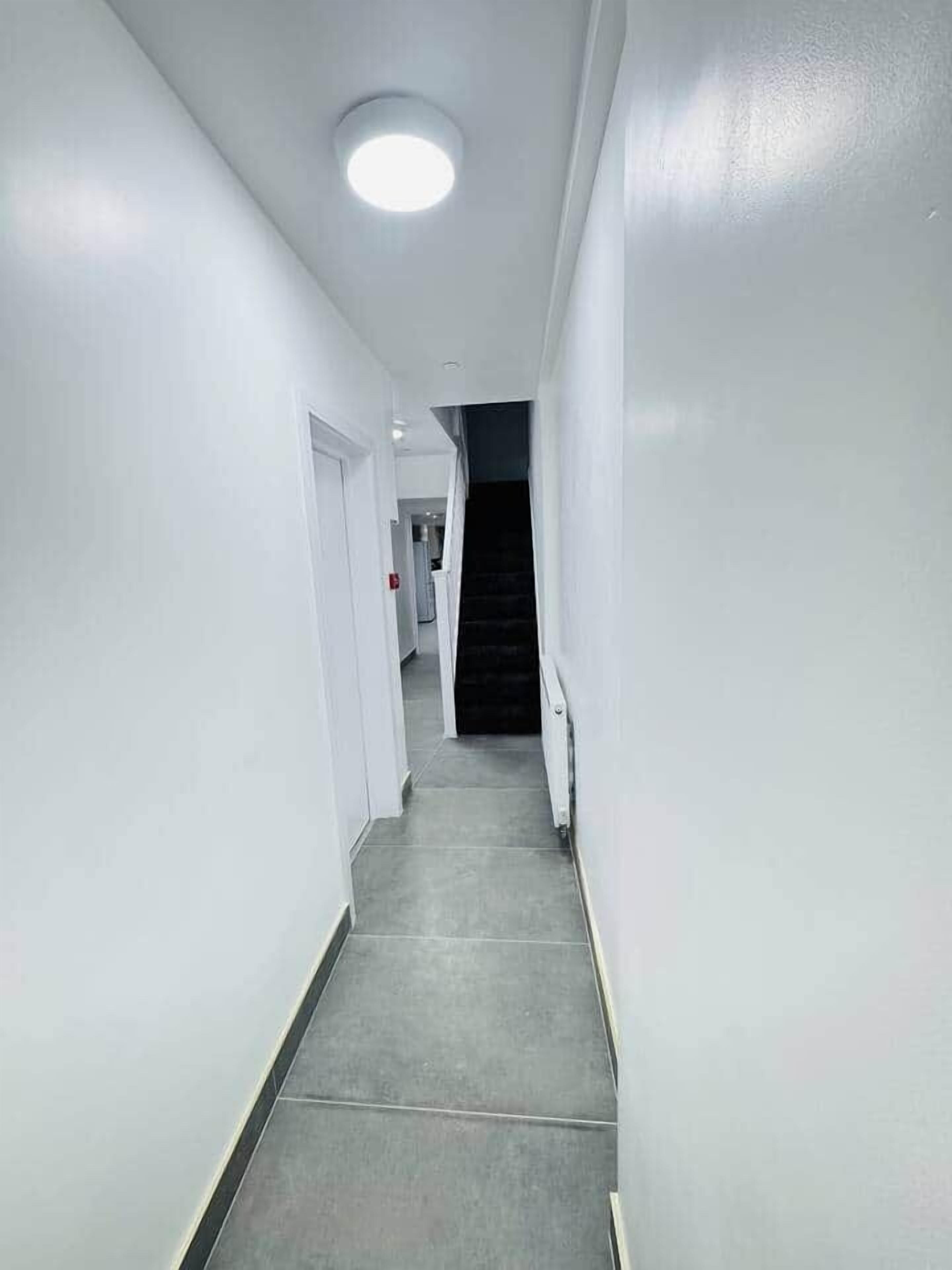 A narrow, well-lit hallway with white walls and a grey floor, leading to a staircase.