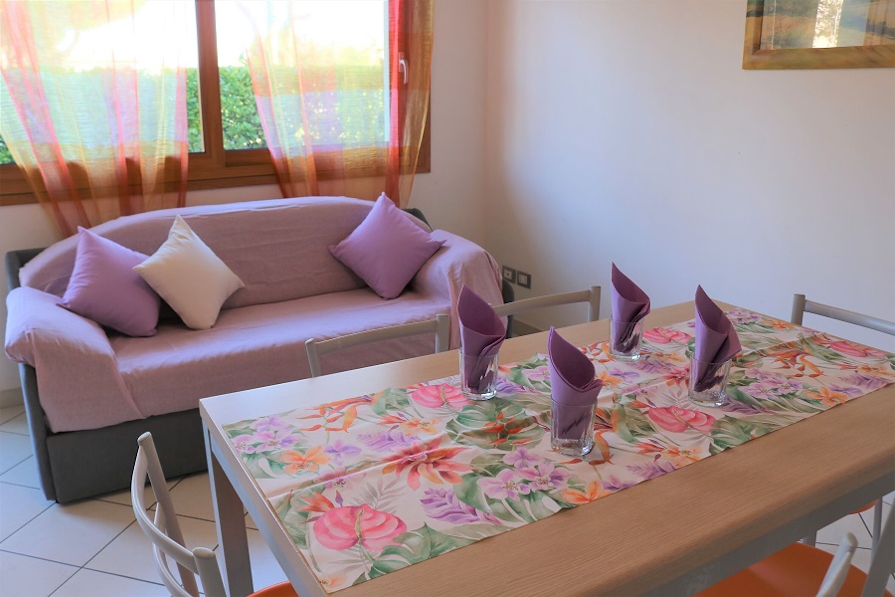 A dining area with a table set for two, a sofa with cushions, and a window with sheer curtains.