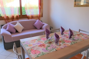 A dining area with a table set for two, a sofa with cushions, and a window with sheer curtains. - Lovely Flat With Patio (San Michele al Tagliamento)