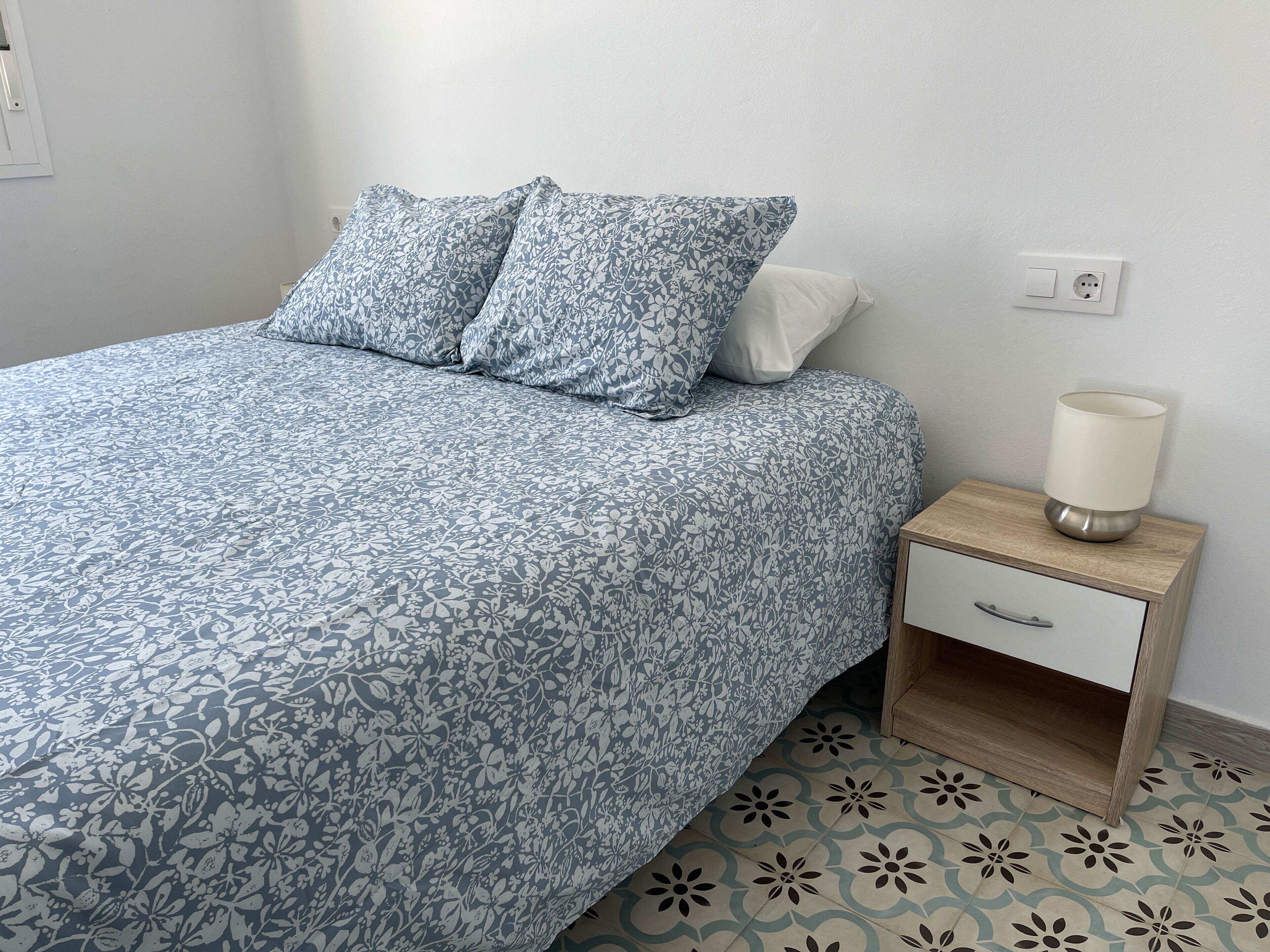 Een slaapkamer met een bed, twee kussens, een nachtkastje met een lamp en een nachtkastje.