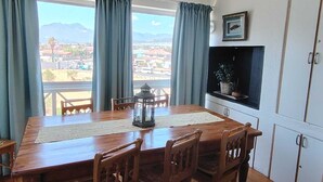 Comedor con mesa de madera, sillas, un farol y vistas a un pueblo a través de amplios ventanales.