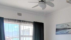 Una habitación de hotel con dos camas individuales, un ventilador de techo, una ventana con cortinas, una mesita de noche con lámpara y un cuadro en la pared.