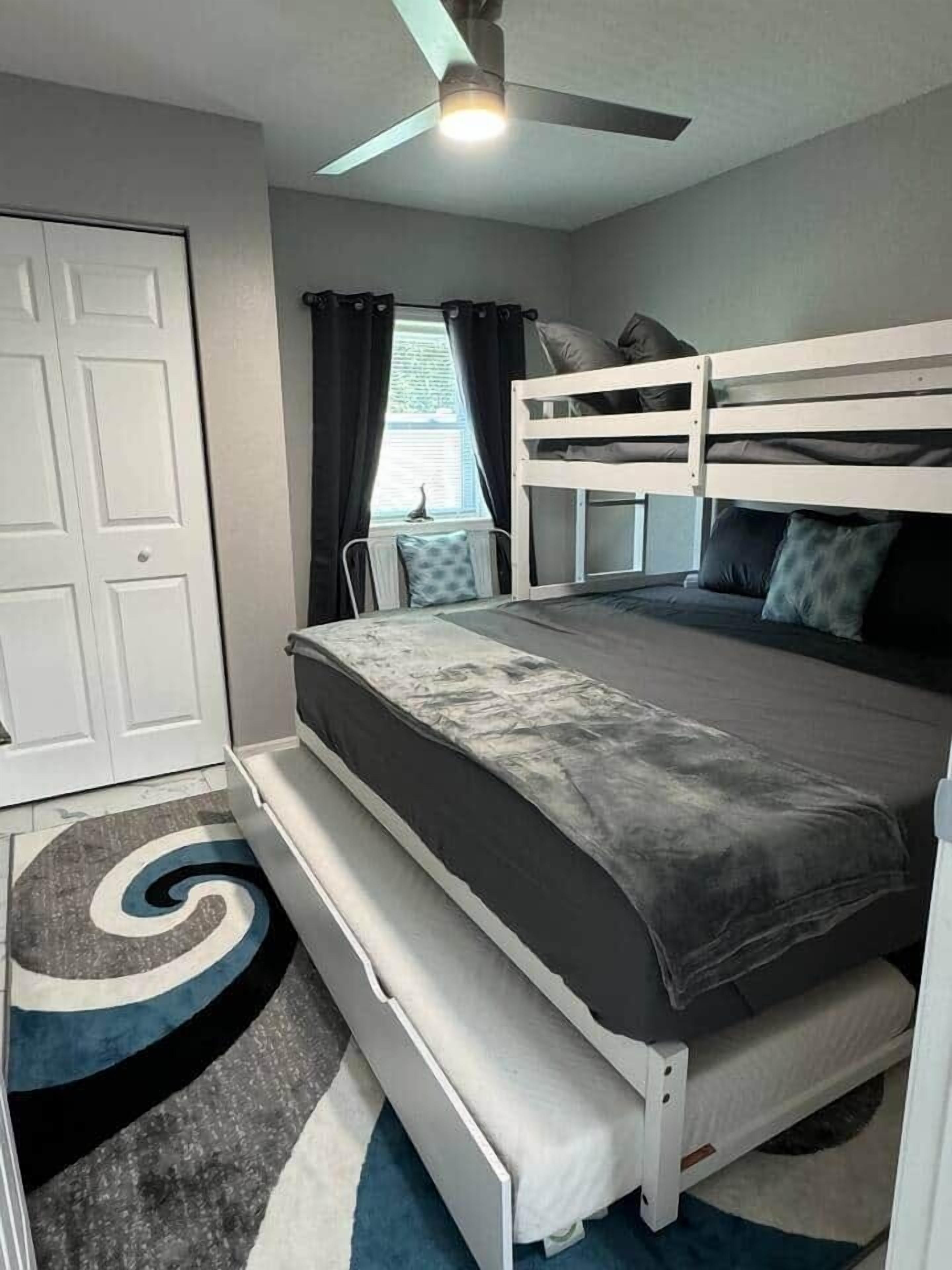 A bedroom with a bunk bed, a ceiling fan, a window with curtains, and a spiral-patterned rug.