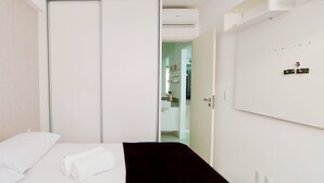 Camera d'albergo con un letto, una coperta nera, cuscini bianchi, una parete bianca, una porta e un piccolo ripiano con oggetti.