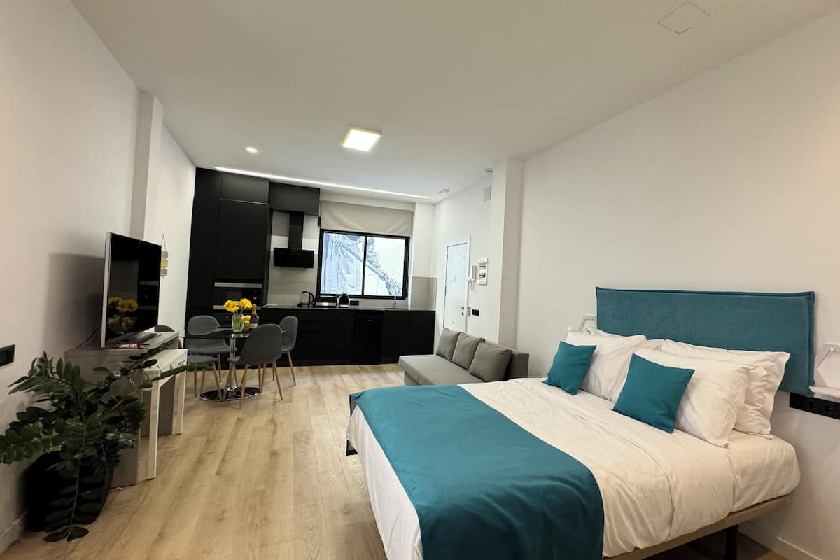 A modern hotel room with a large bed, a desk with a chair, a kitchenette, and a view of the city.