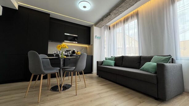 Una sala moderna con un sofá gris, dos sillas grises, una mesa de comedor con un florero de flores amarillas y una cocina al fondo.