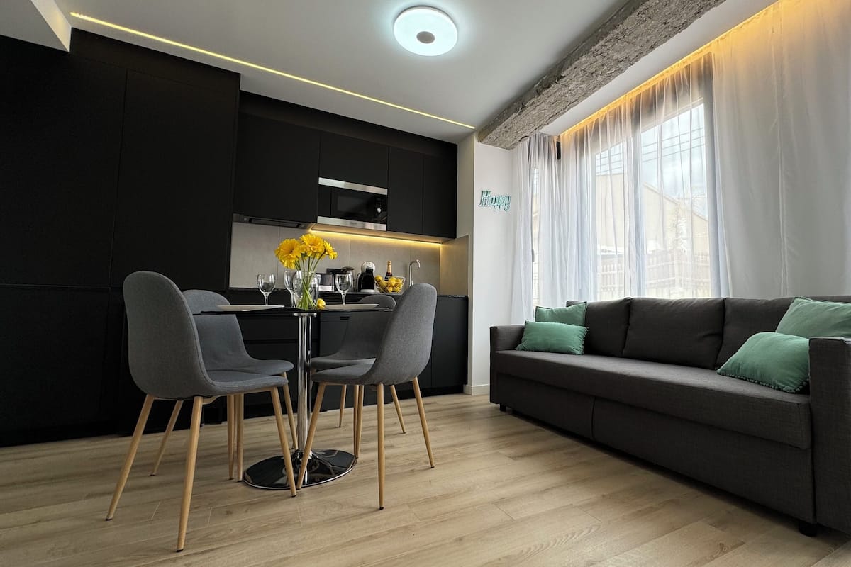 A modern living room with a grey sofa, two grey chairs, a dining table with a vase of yellow flowers, and a kitchen in the background.