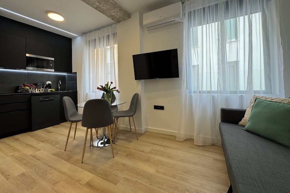 A modern living room with a dining area, a flat-screen TV mounted on the wall, and a kitchen area with a microwave and sink.