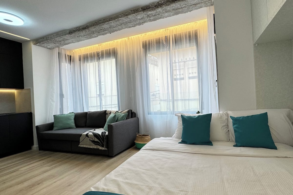 A hotel room with a bed, a sofa, and a window with sheer curtains.