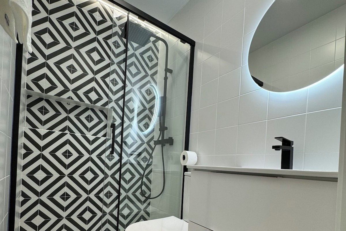 A bathroom with a geometric patterned floor, a white toilet, a glass-enclosed shower with a black hose, and a wall-mounted mirror.