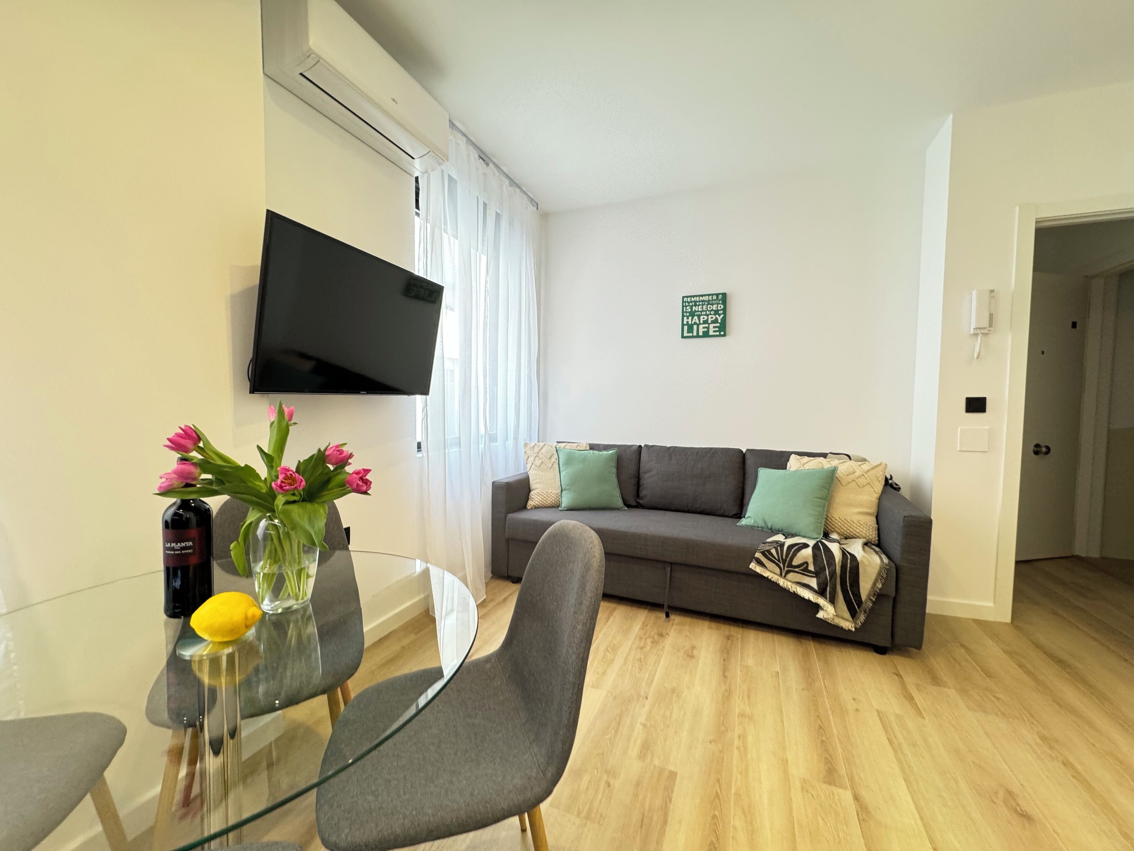 A modern living room with a flat-screen TV, a glass table, and a grey sofa with patterned cushions.