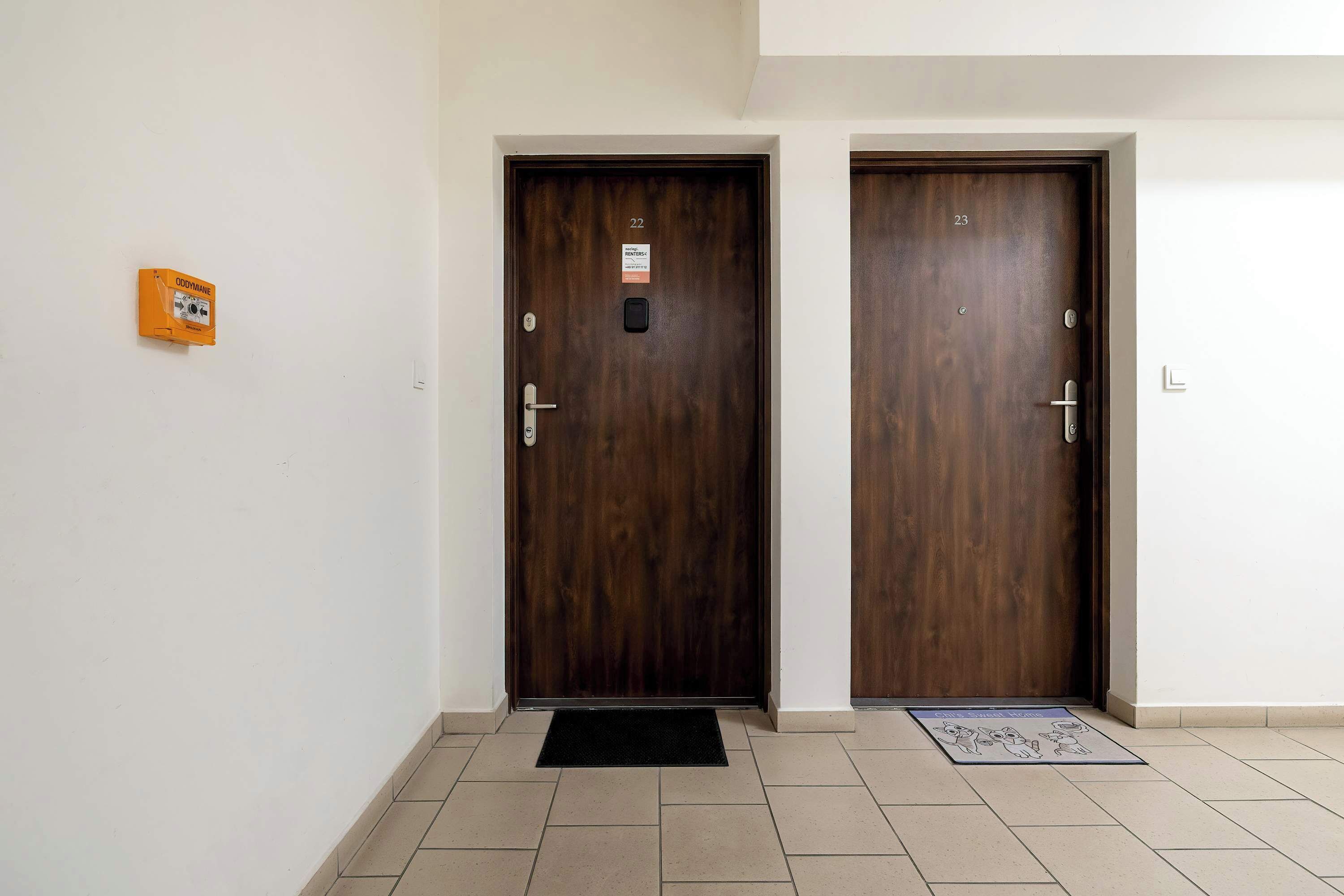 Dos puertas de madera con letreros numerados, un pequeño dispositivo naranja en la pared y un felpudo negro en la entrada.