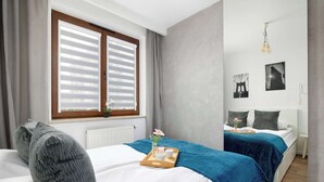 Hotellihuone, jossa on kaksi sänkyä, ikkuna, jossa on sälekaihtimet, tarjotinalusta, jossa on kynttilä ja kukka, sekä seinällä olevia kehystettyjä kuvia.