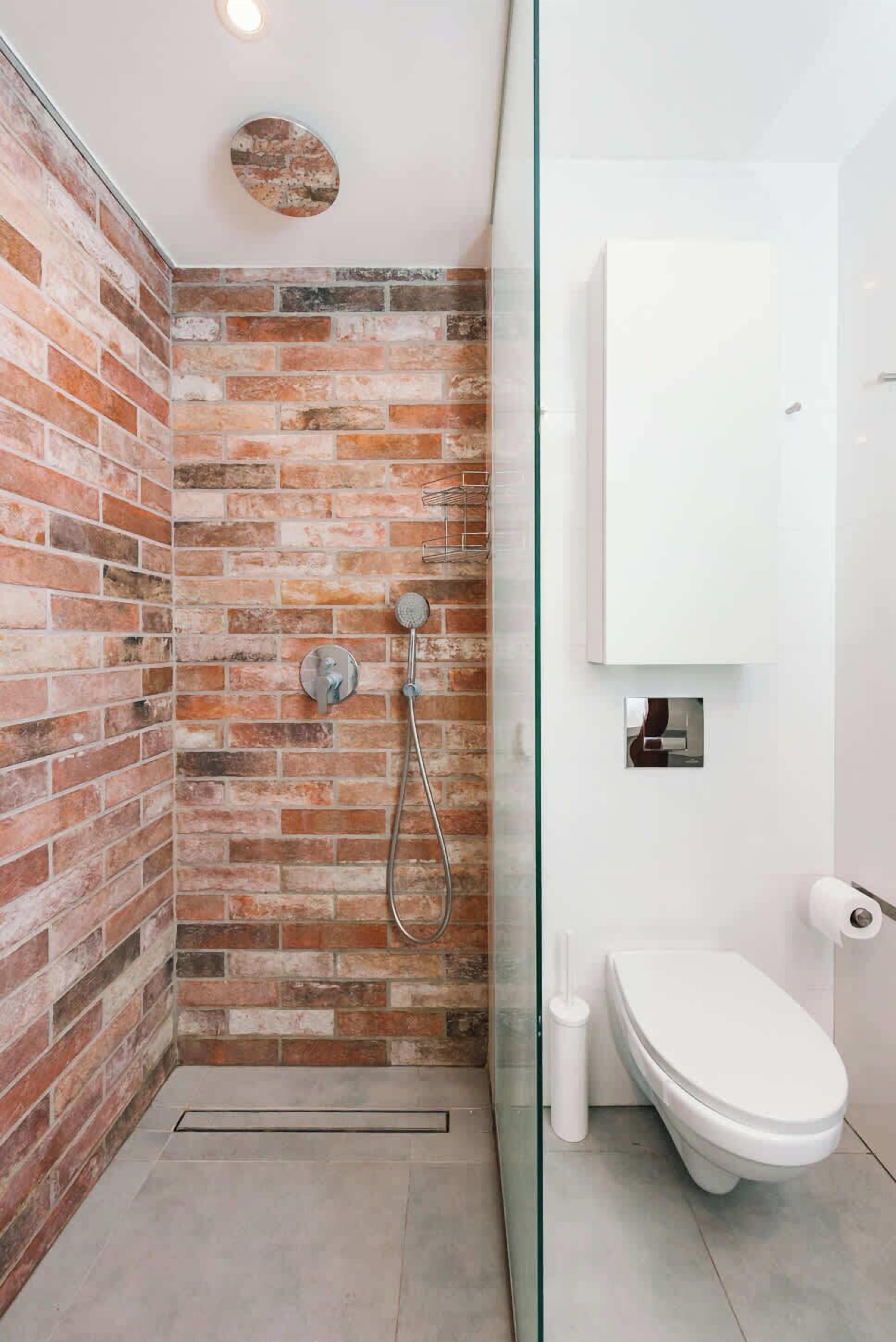 A bathroom with a brick wall, a glass shower enclosure, a white toilet, and a white sink.