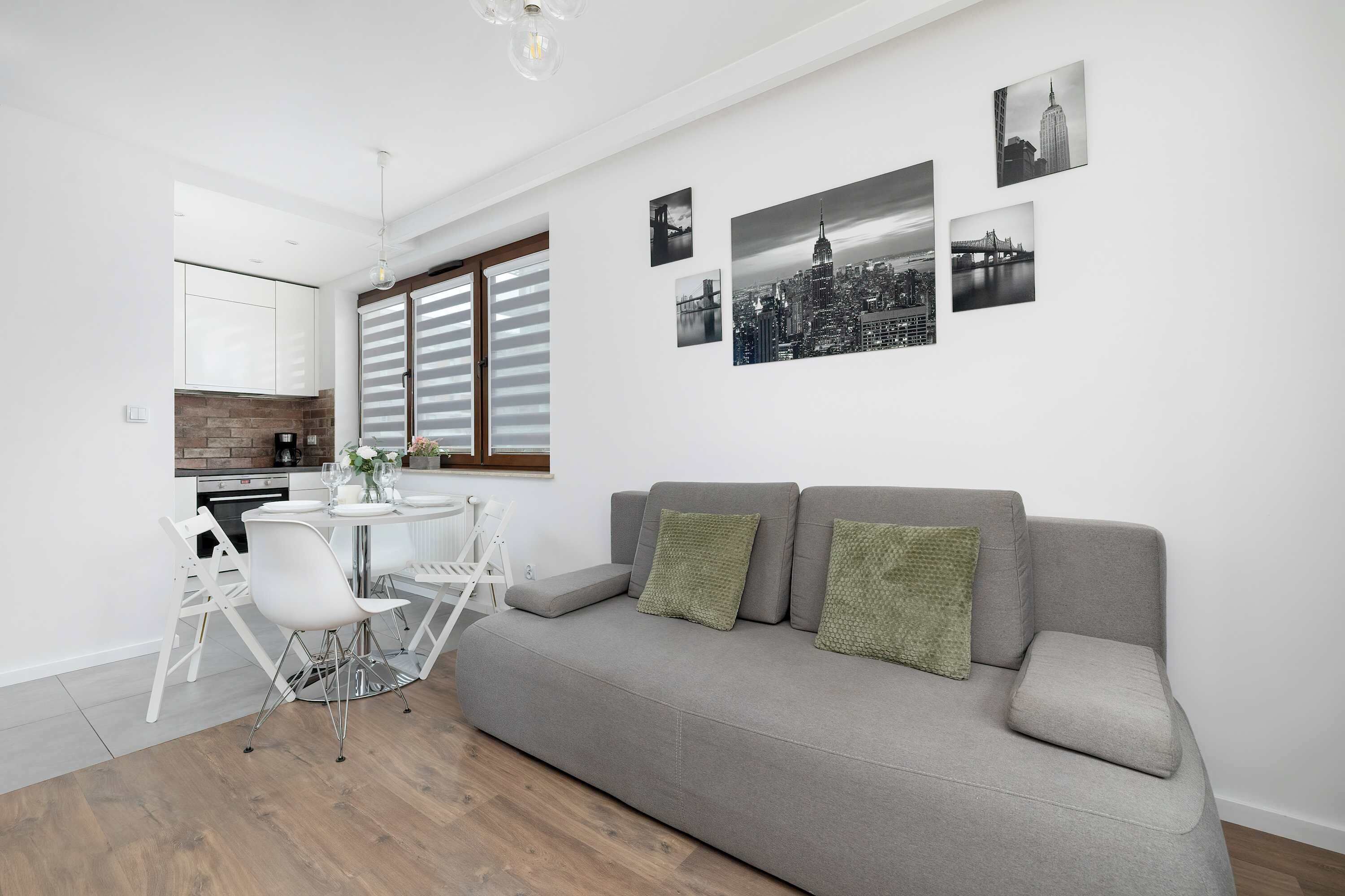 A modern living room with a grey sofa, white dining chairs, and a small round table. There are black and white cityscape photographs on the wall.