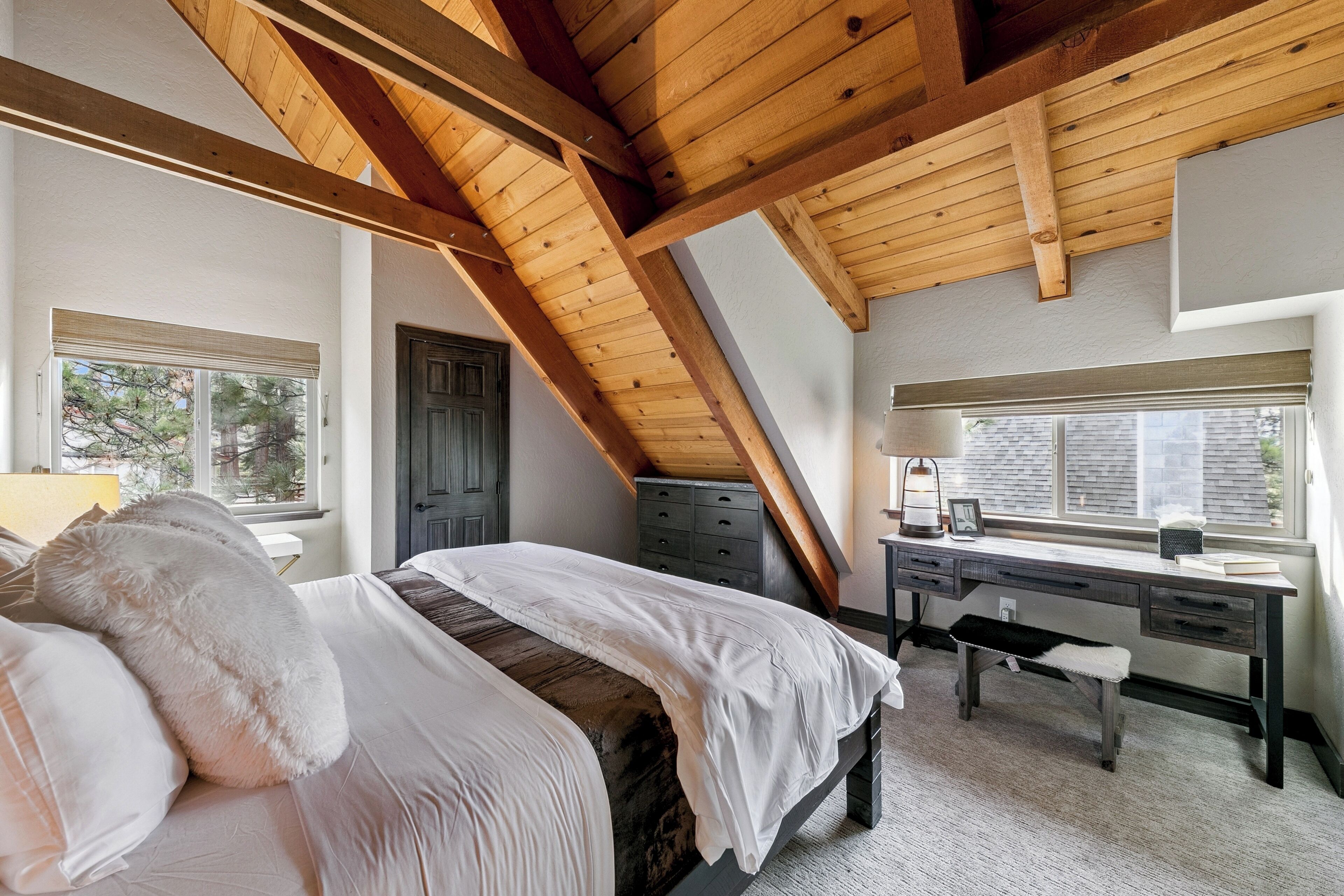 A bedroom with a wooden ceiling, a bed with white bedding, a desk, and a window with blinds.
