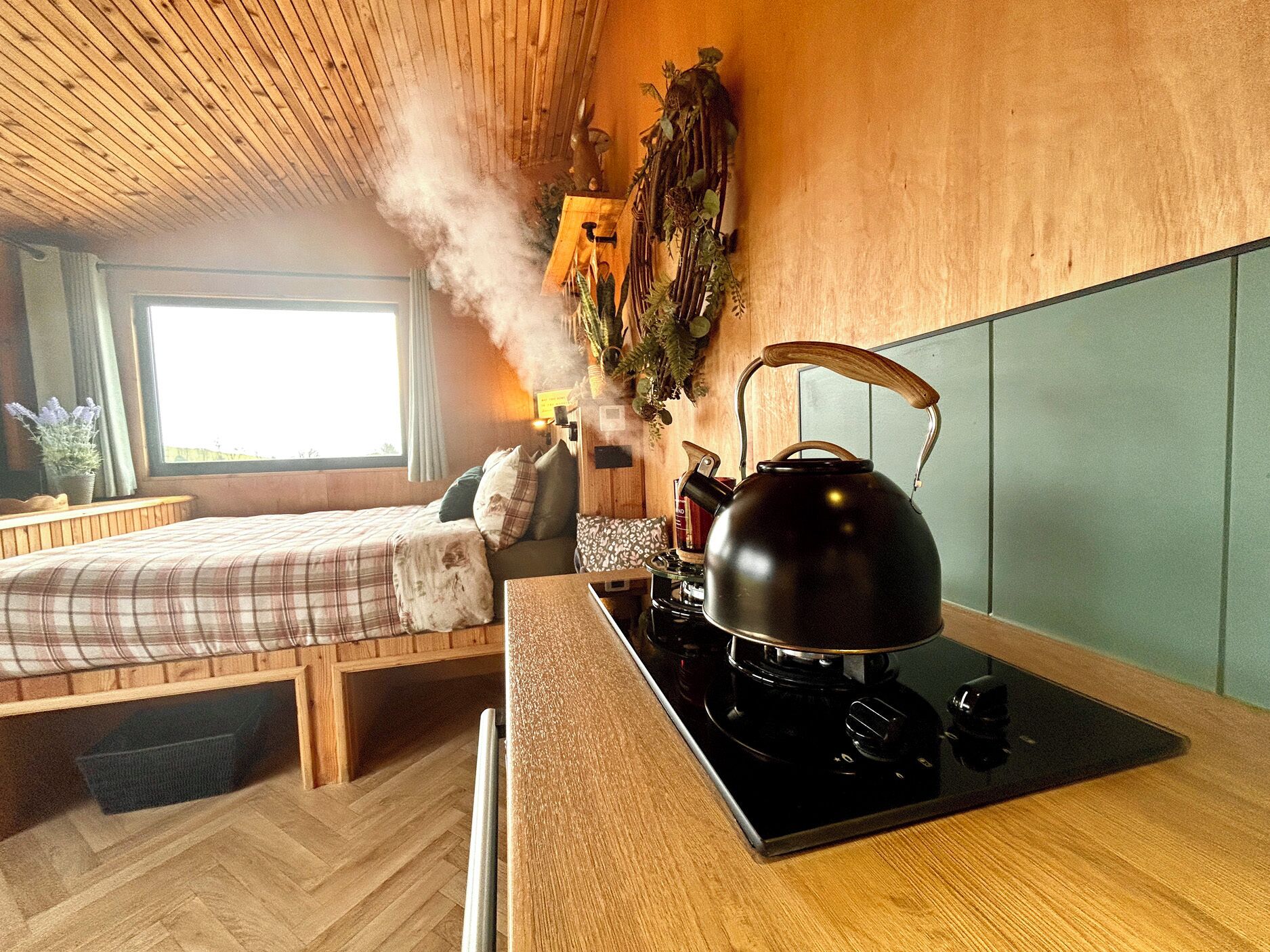 A cozy room with a bed, a wooden ceiling, a window, a kettle, and a wall-mounted plant.
