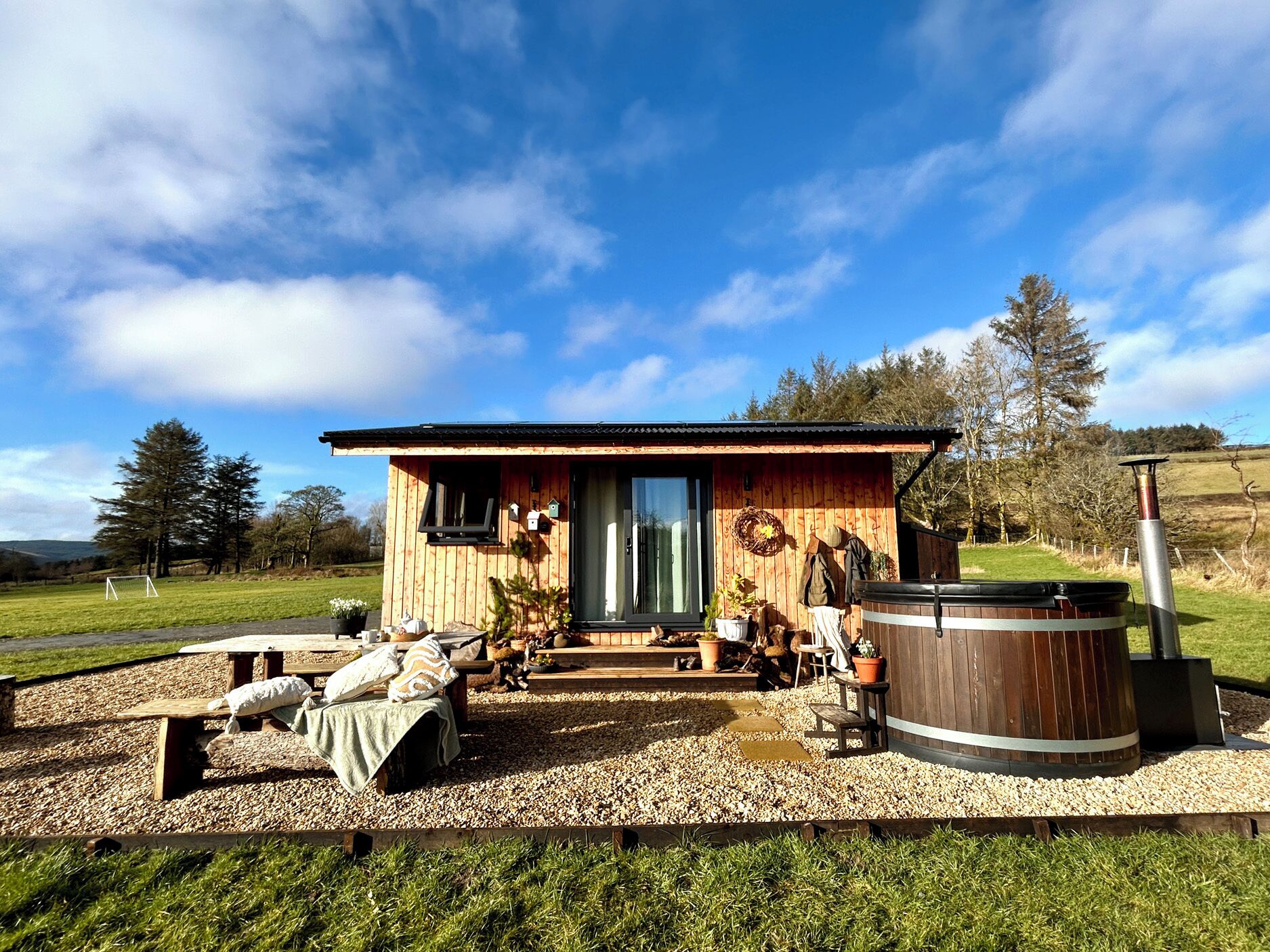 A wooden cabin with a large hot tub, a patio with a table and benches, and a view of the surrounding landscape.