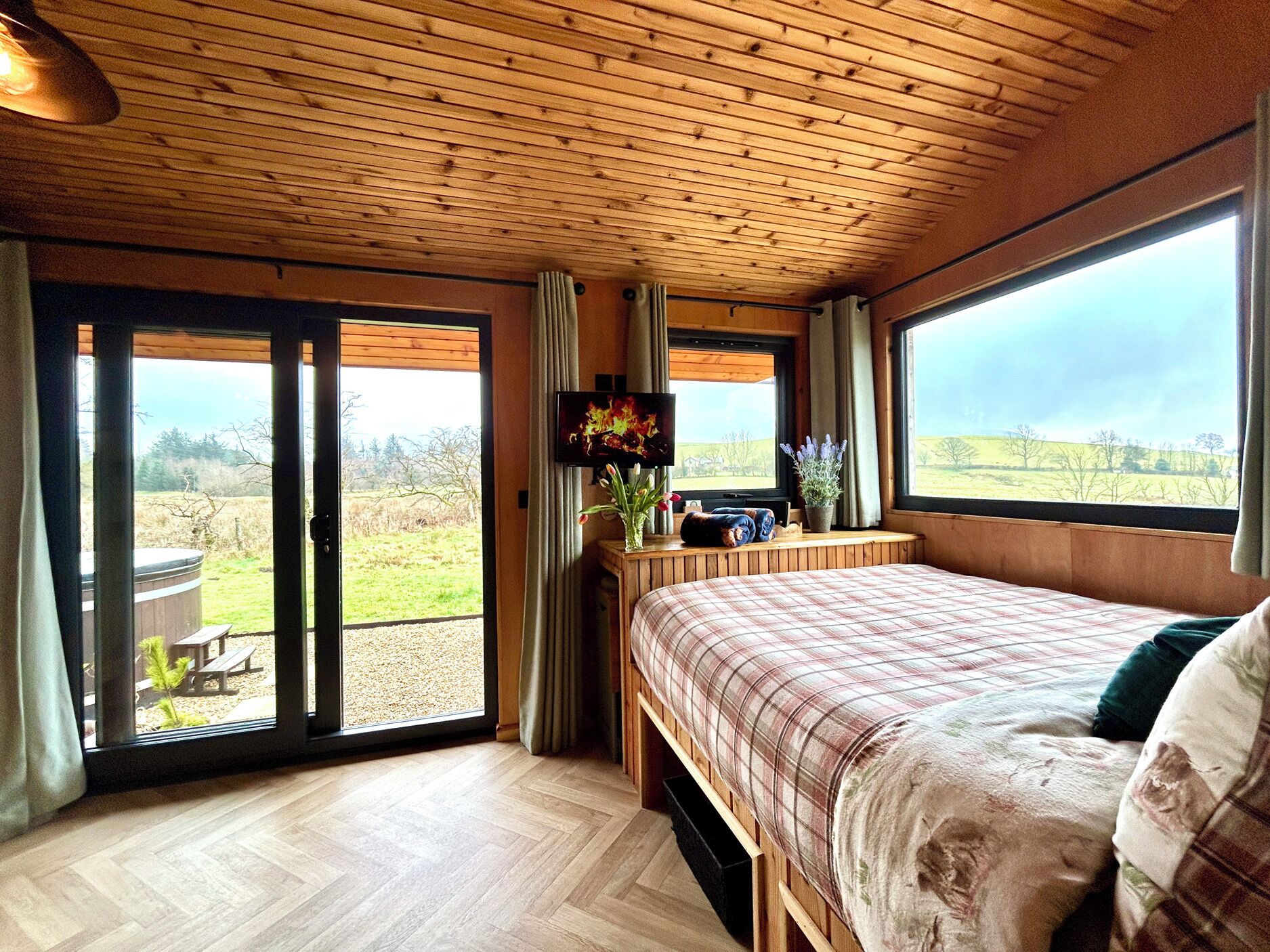 A bedroom with a bed, a TV, a window with a view of the outdoors, and a hot tub visible through the sliding doors.
