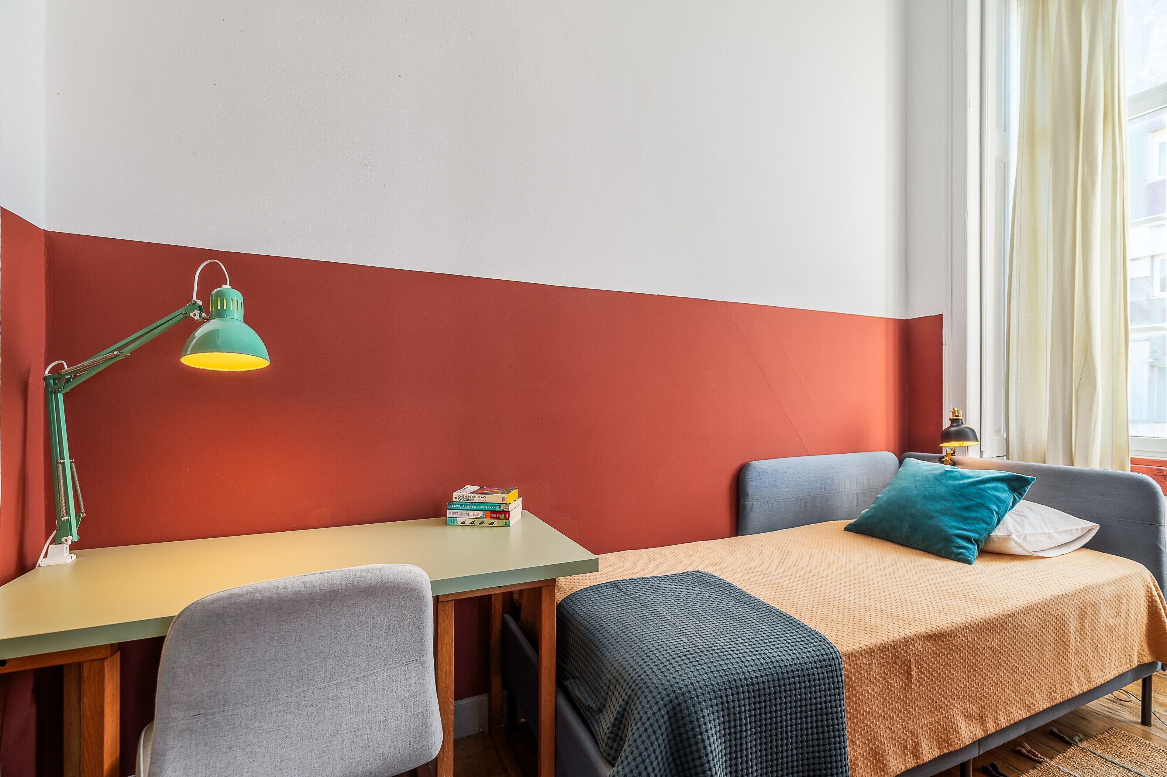 Ein Zimmer mit Schreibtisch, zwei Stühlen, einem Bett mit blauer Decke, einer grünen Schreibtischlampe und einem Fenster mit Vorhängen.