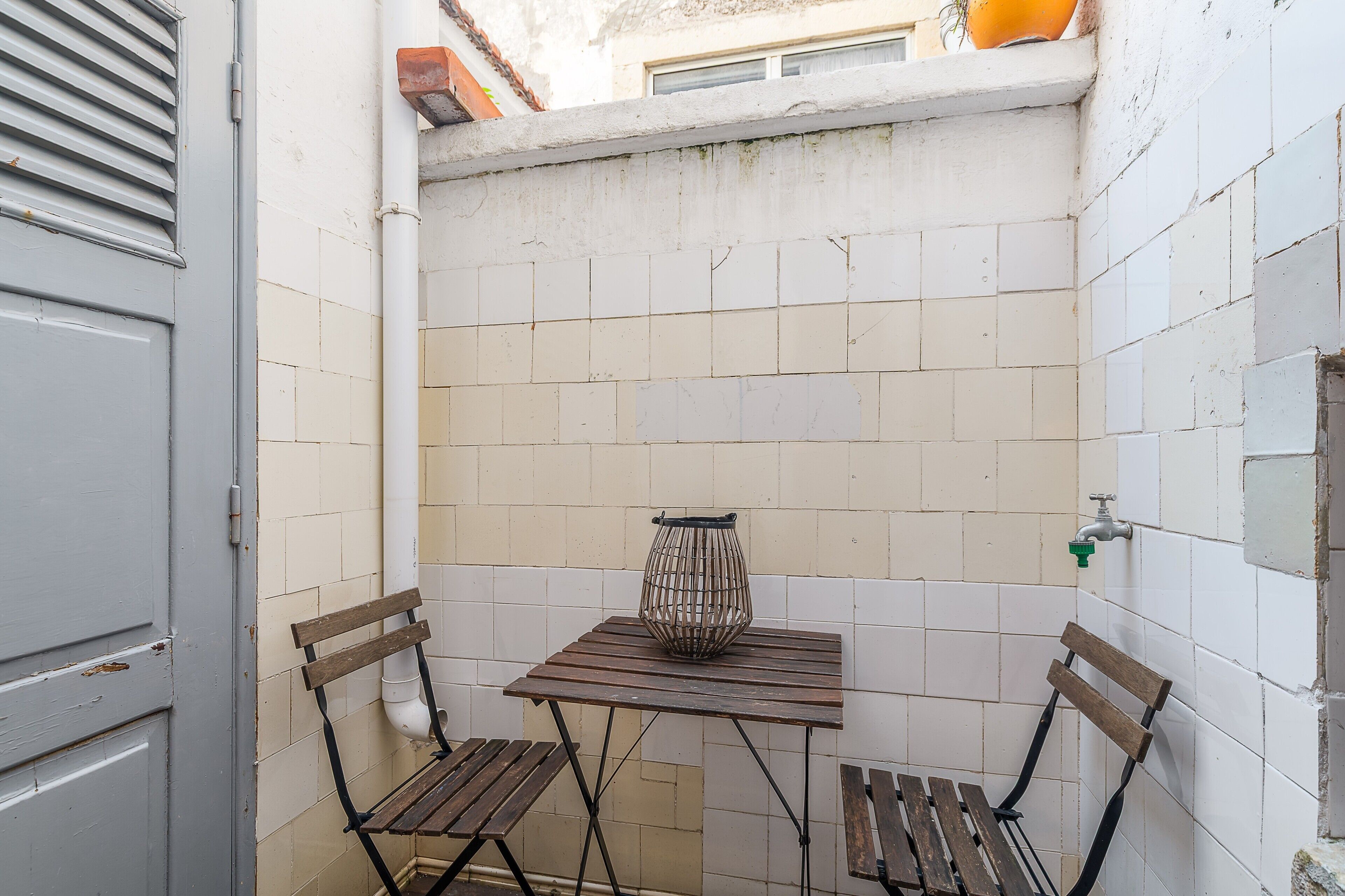 Um pequeno quarto com piso de azulejos, uma mesa e duas cadeiras, um vaso de planta e uma janela.