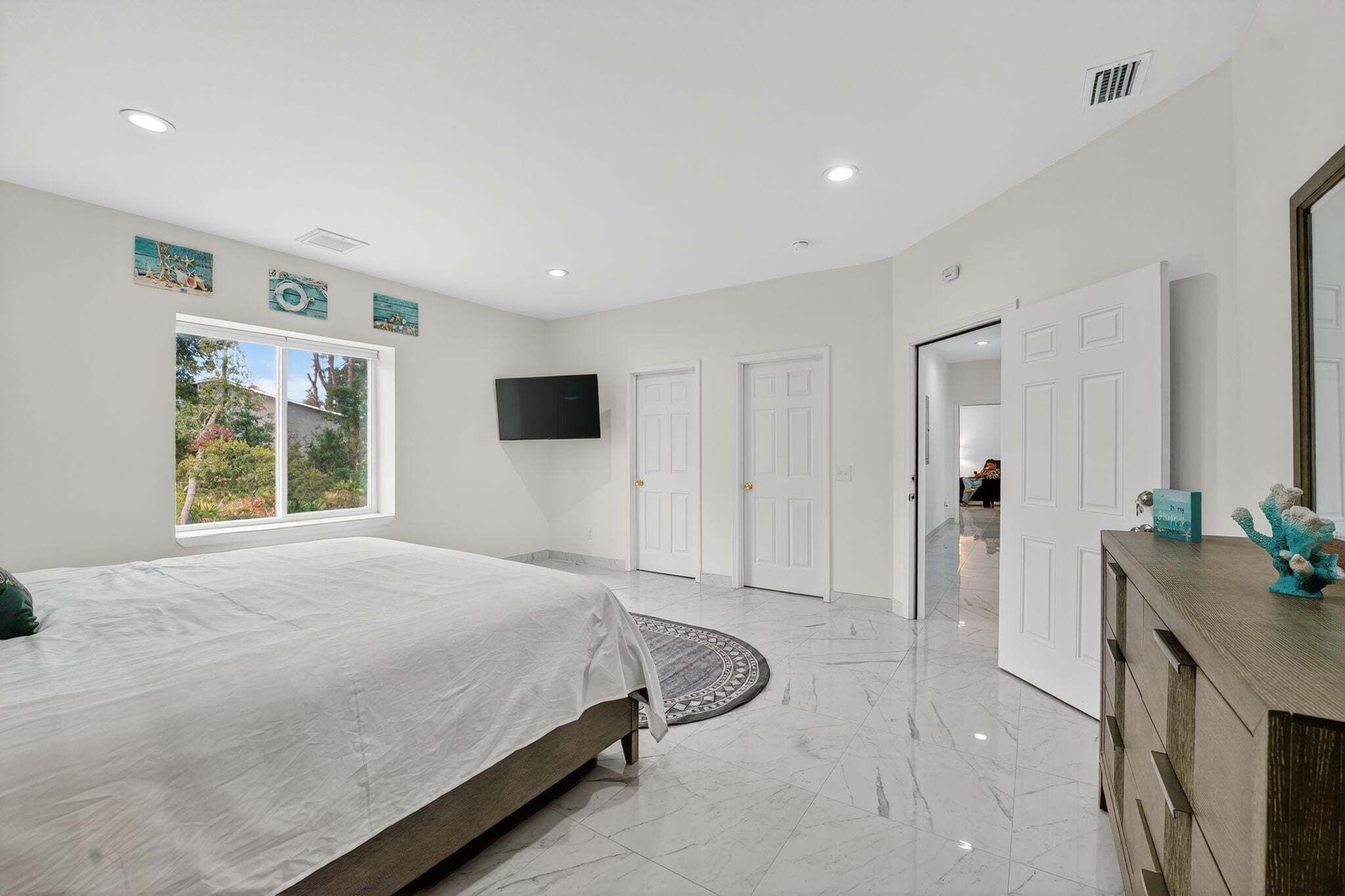 A modern bedroom with a large bed, a TV mounted on the wall, and a view of the outdoors through a window.