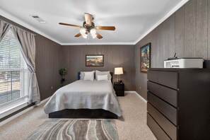 Une chambre à coucher comprenant un lit, un ventilateur de plafond, une table de chevet, une commode et une fenêtre avec des rideaux.