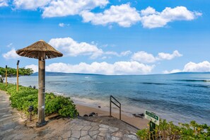 A beach with a thatched umbrella, a caution sign, and a stone pathway leading to the water. - SUMMER SALE! Ocean-view 1BR retreat with resort comforts in Lahaina, Kaanapali Shores 544 (Lahaina)