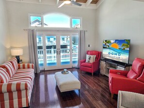 Ein Wohnzimmer mit einem gestreiften Sofa, einem roten Sessel, Holzboden, einem Fernseher und einem Balkon mit Meerblick.