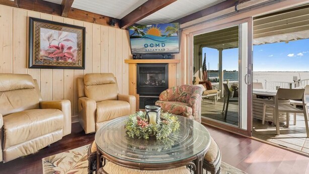 Ein gemütliches Wohnzimmer mit Ledersesseln, einem Glastisch mit floralem Mittelstück, einem Kamin und einem Fernseher, der 'OCMD GETAWAYS' anzeigt.