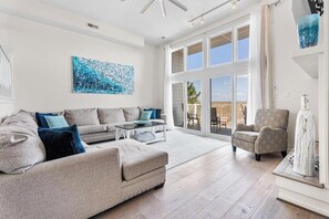 Ein geräumiges Wohnzimmer mit einer großen Eckcouch, einem Couchtisch und einem Sessel. Es gibt ein großes Fenster mit Blick ins Freie.