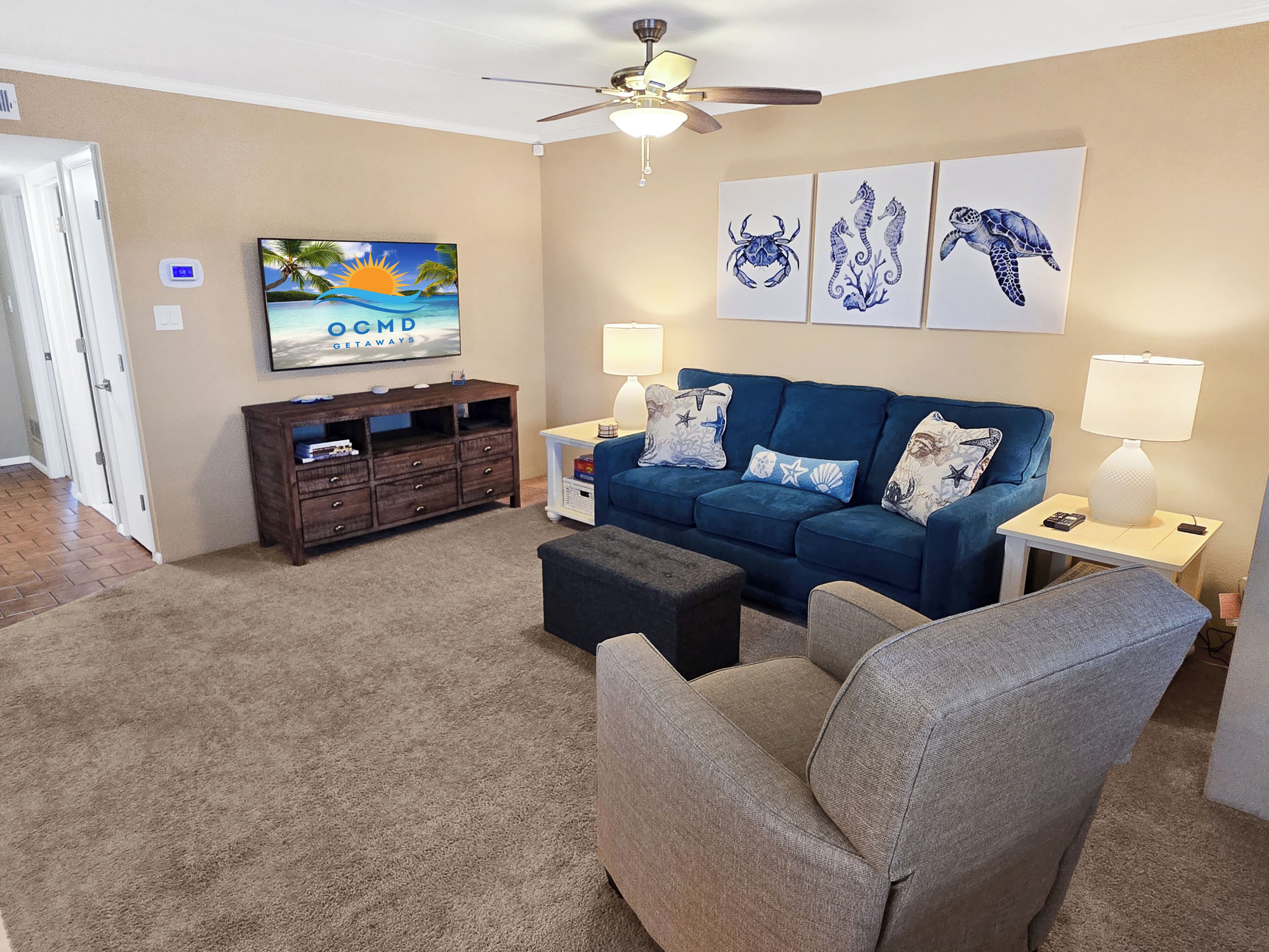 Uma sala de estar com um sofá azul, duas mesas laterais com abajures, uma mesa de centro, uma TV sobre um suporte de madeira e um ventilador de teto.