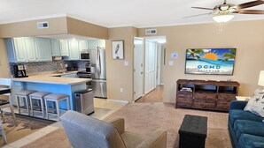 Een woonkamer met een grijze fauteuil, een blauwe bank en een televisie waarop 'OCMD GETAWAYS' staat. Op de achtergrond is een keuken met lichtblauwe keukenkastjes en een koelkast te zien.