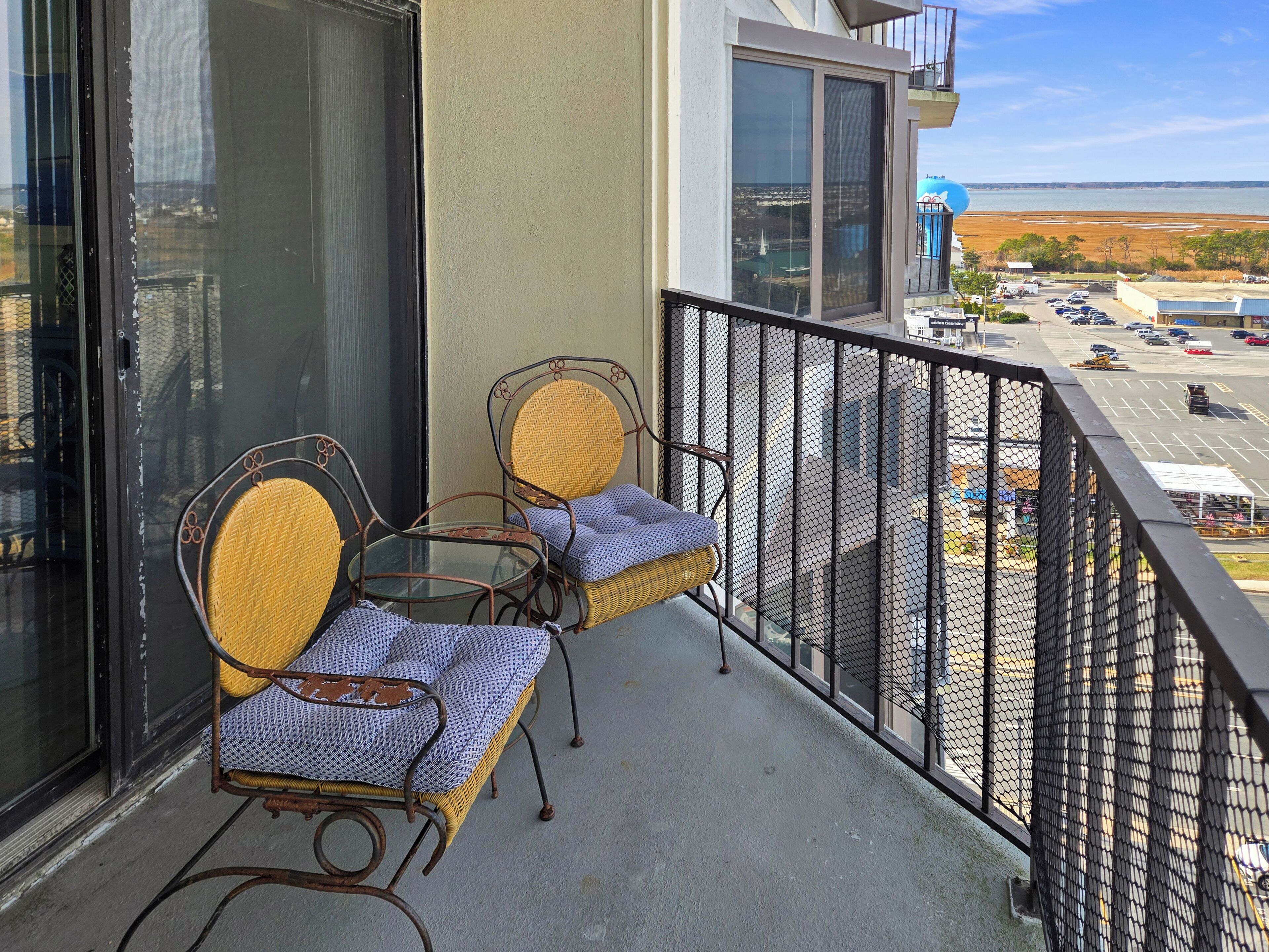 A balcony with two wicker chairs and a glass table, overlooking a parking lot and a body of water.