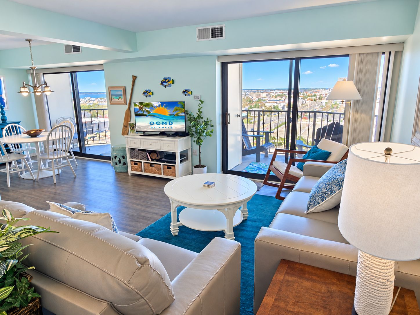A spacious living room with a balcony, a dining area, and a television displaying 'OCMD GETAWAYS'.