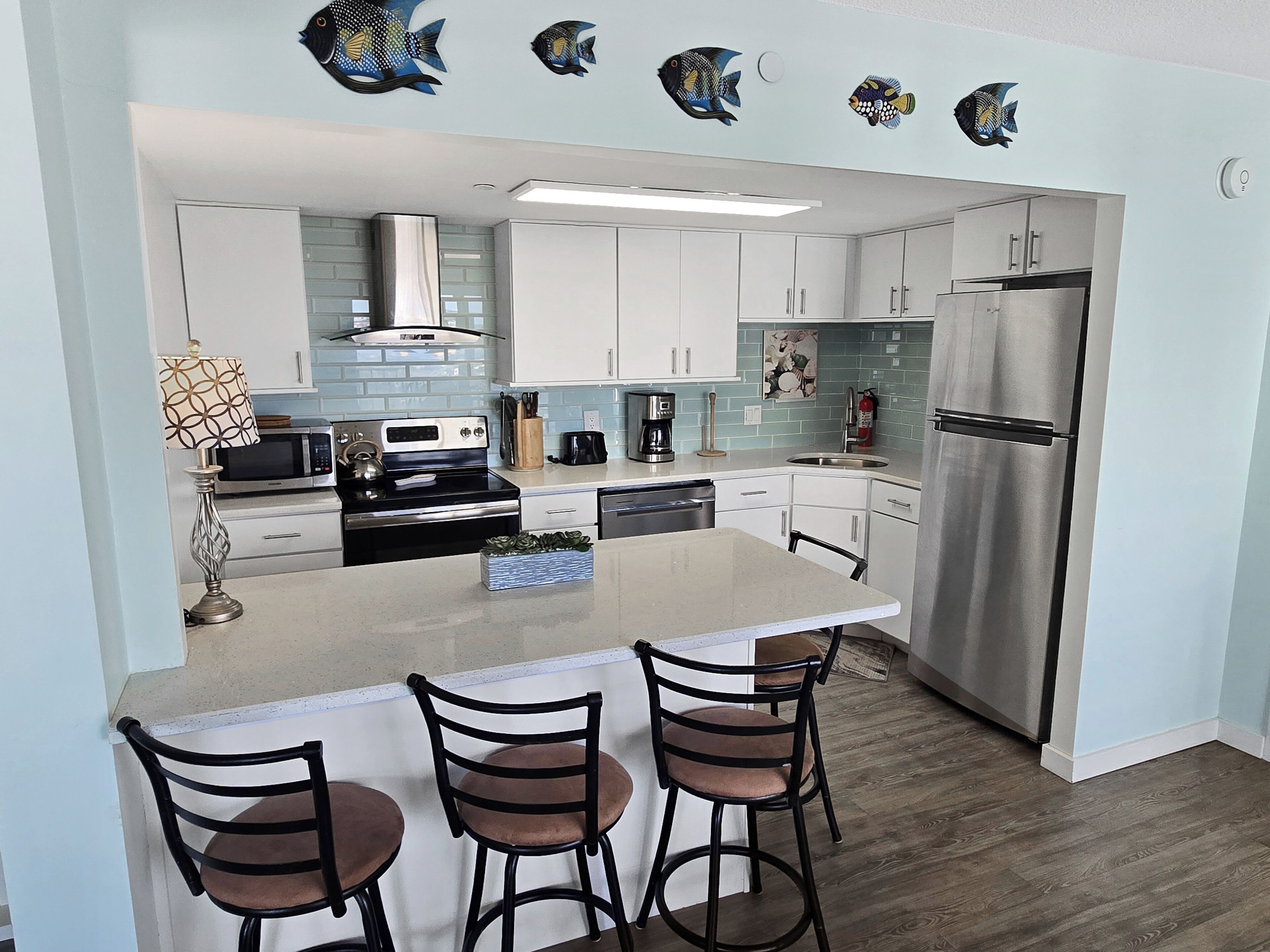 A modern kitchen with white cabinets, a stainless steel refrigerator, a central island with bar stools, and decorative fish wall art.