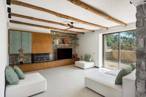 Ein modernes Wohnzimmer mit Kamin aus Stein, Holzbalken und einem großen Fenster mit Blick auf eine Steintreppe.