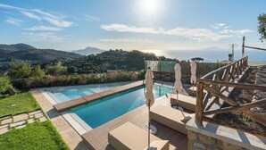 Una piscina con bordo in legno e lettini prendisole, circondata da un muro in pietra e affacciata sul mare e sulle colline.