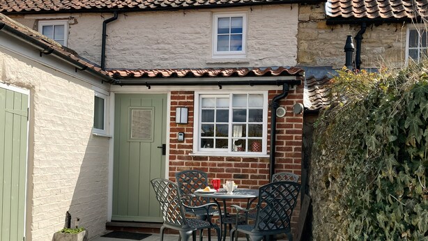 A small patio with a table and chairs, a green door, and a window with a brick wall. - Beacon Cottage (Pickering)