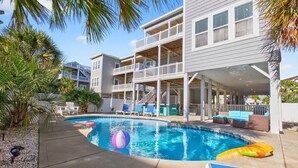 A pool area with a multi-level building, lounge chairs, and palm trees. - Sea Esta is a 7-bedroom, 5-bathroom, large enough for the whole family! (Ocean Isle Beach)