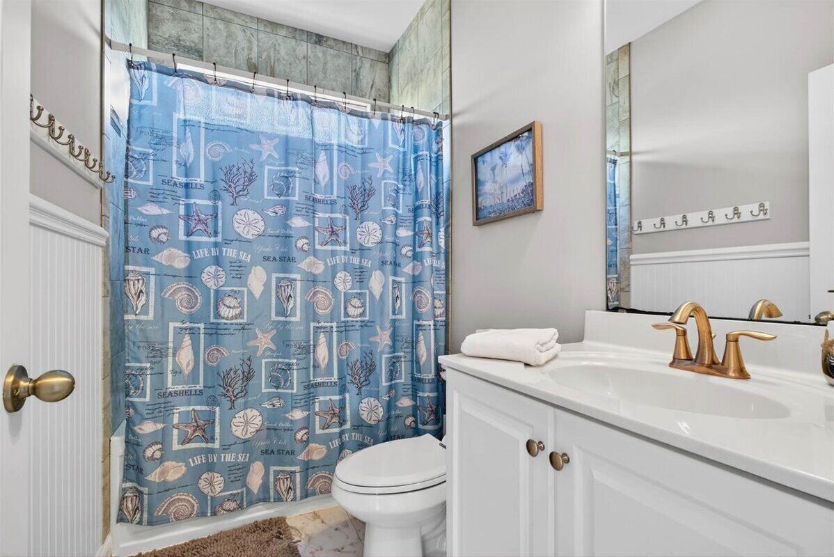 A bathroom with a blue seashore-themed shower curtain, white toilet, and a white sink with gold faucets.