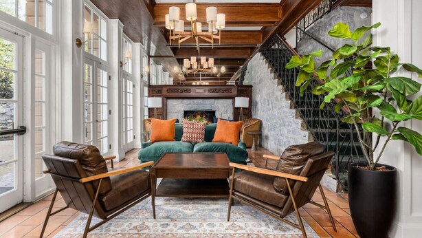 A spacious living room with a large sofa, two armchairs, a coffee table, and a fireplace. There is a staircase with a metal railing on the right and a large window on the left.