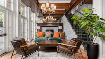 A spacious living room with a large sofa, two armchairs, a coffee table, and a fireplace. There is a staircase with a metal railing on the right and a large window on the left.