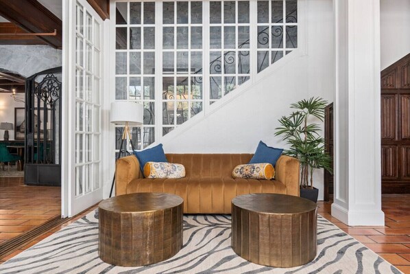 A living room with a brown sofa, two round wooden side tables, a zebra-patterned rug, and a staircase with a white railing.