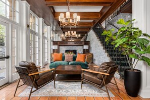 A spacious living room with a large sofa, two armchairs, a coffee table, and a fireplace. There is a staircase with a metal railing on the right and a large window on the left.