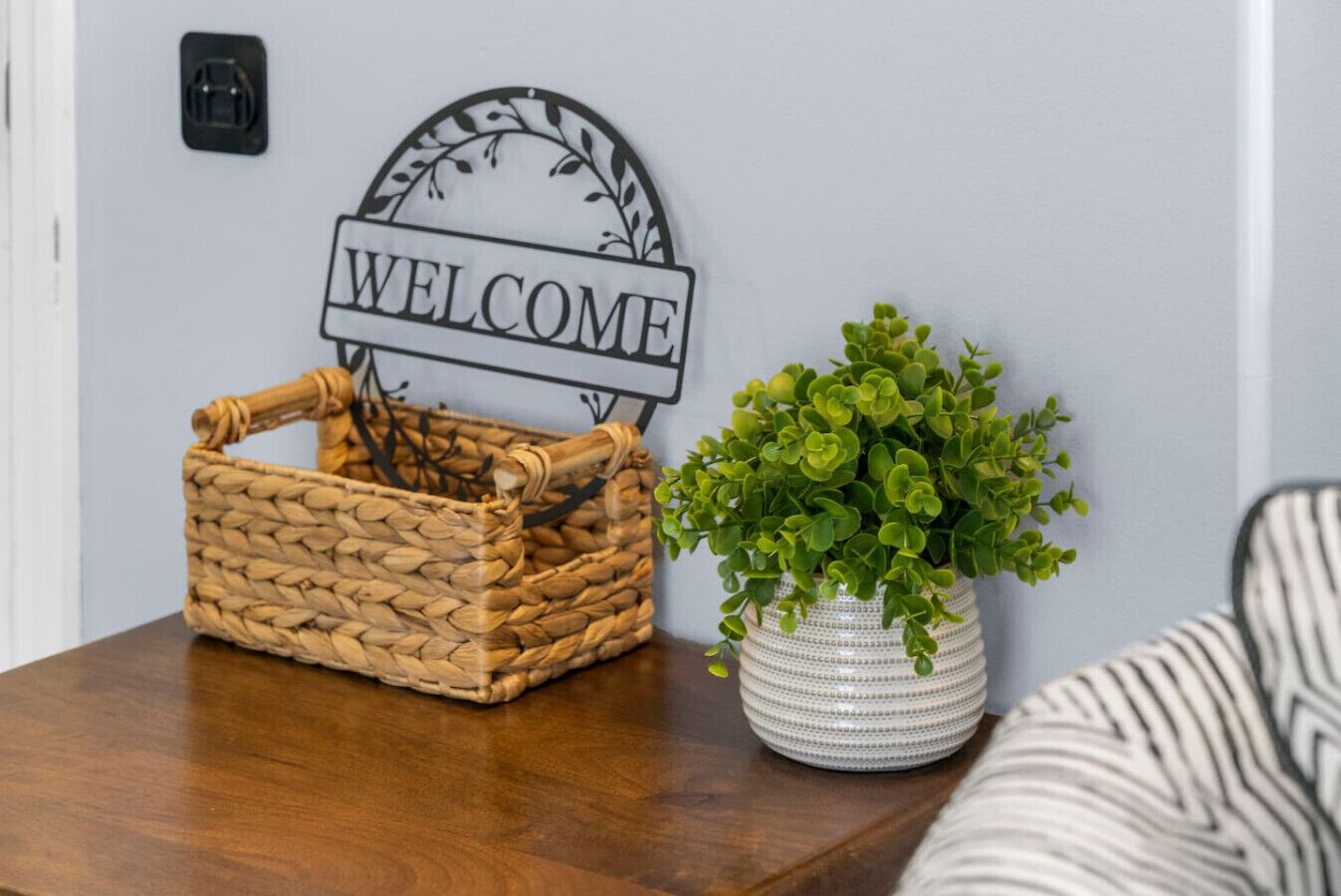 A wicker basket and a potted plant on a wooden table with a 'WELCOME' sign above.
