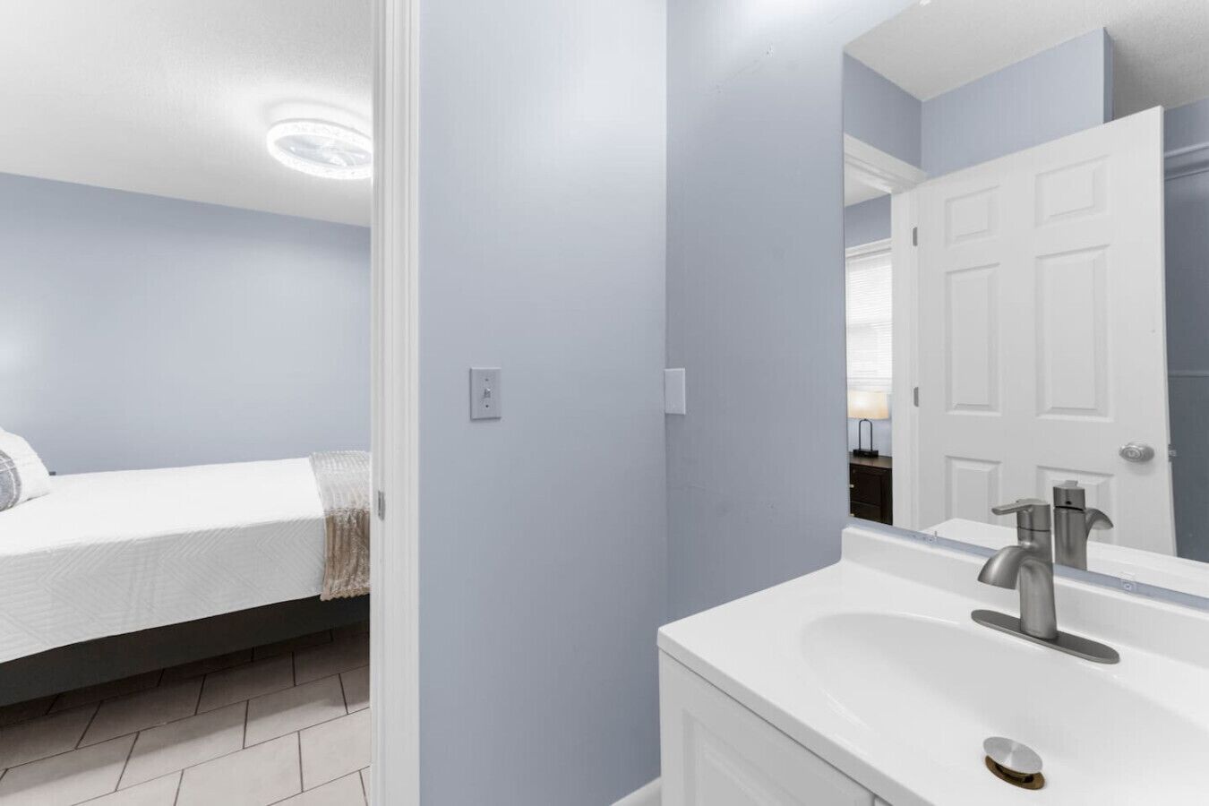 Une chambre moderne avec un lit, une salle de bain équipée d’un lavabo et d’un miroir, et une porte donnant sur une autre pièce.