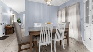 Een eetgedeelte met een houten tafel, witte stoelen, een kroonluchter en lichtblauwe muren.