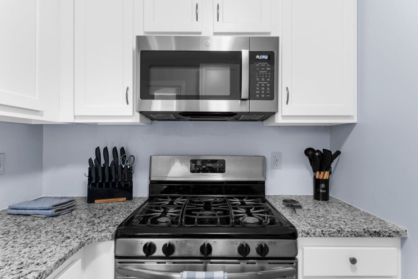 Una cucina con mobili bianchi, un forno a microonde in acciaio inox, un fornello a gas e set di coltelli sul piano di lavoro.