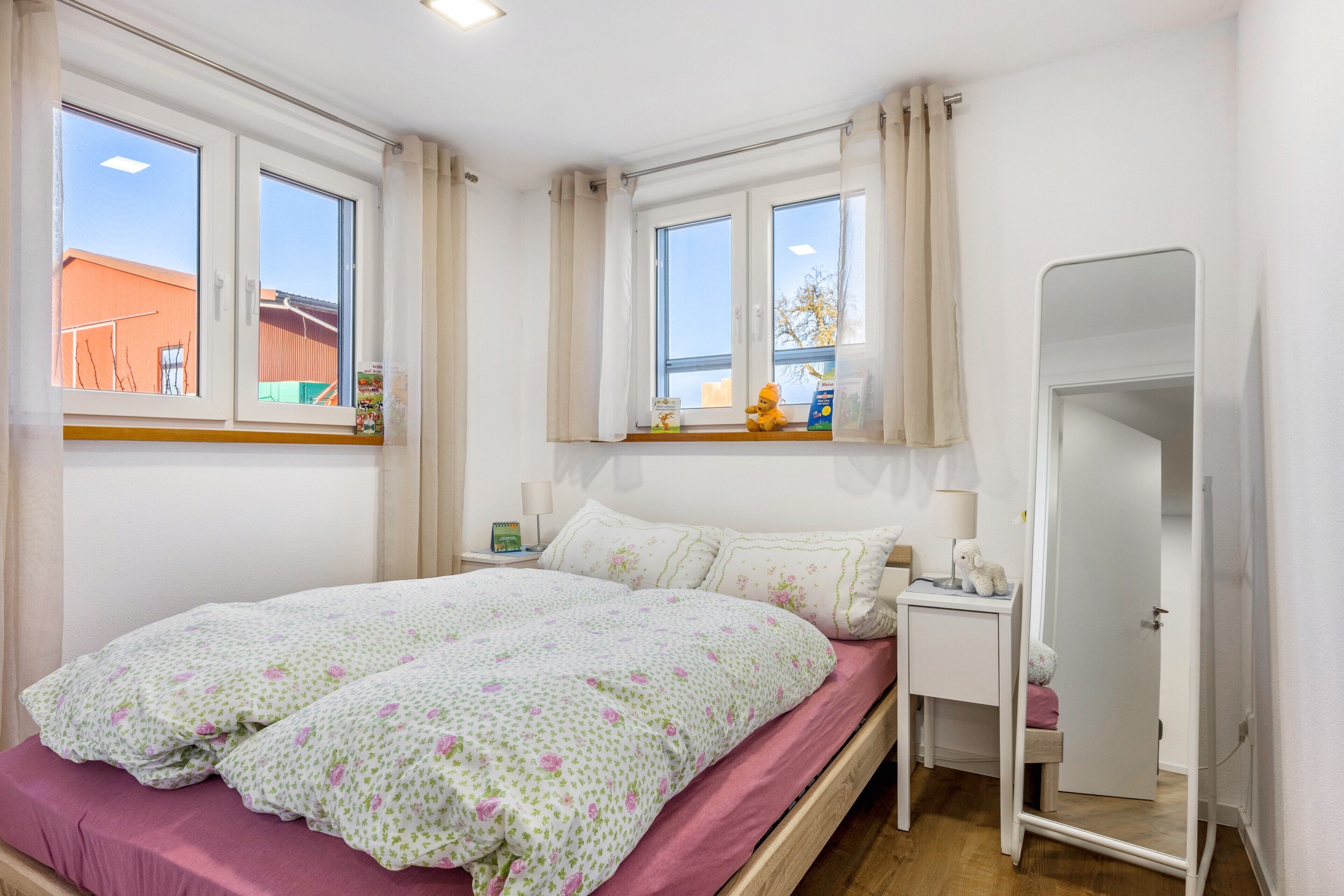 Un dormitorio con cama, una mesita de noche con lámpara, un espejo y una ventana con cortinas.